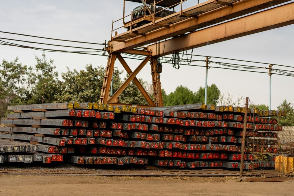 Industrial steel beams stacked outdoors beneath a gantry crane, with a backdrop of trees.
